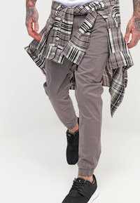 Man met getatoeëerde handen, gekleed in grijze joggingsbroek, zwarte sneakers en een geruit overhemd om de taille geknoopt, die naar voren loopt.