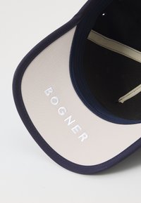 Casquette bleu marine avec un dessous beige. Présente "BOGNER" brodé en blanc. Matériau lisse avec une forme structurée et une visière incurvée.