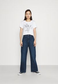 T-shirt branca com gráfico floral preto e texto "Levi's", combinada com jeans de perna larga azul-escuro e sapatilhas de lona pretas.