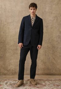 Blazer navy con due tasche frontali, abbinato a una camicia beige a fantasia. Pantaloni scuri e scarpe in suede color tan completano il look.