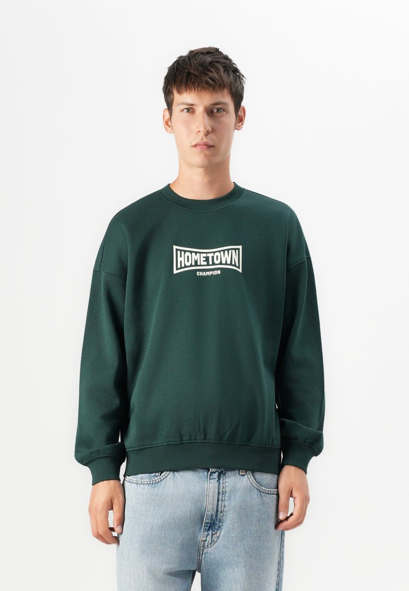Young man wearing dark green sweatshirt with "HOMETOWN CHAMPION" text and light blue jeans, standing against white background.