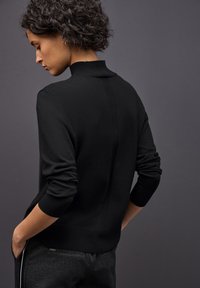 Pull en tricot noir avec un col montant, doté de poignets et d'ourlet côtelés, et d'un détail de couture verticale descendant le dos.