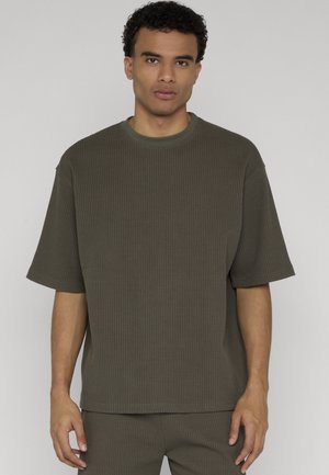 Bărbat cu păr scurt creț, purtând o cămașă largă kaki cu model waffle și mâneci scurte, asortată cu pantaloni, stând în picioare pe un fundal simplu.