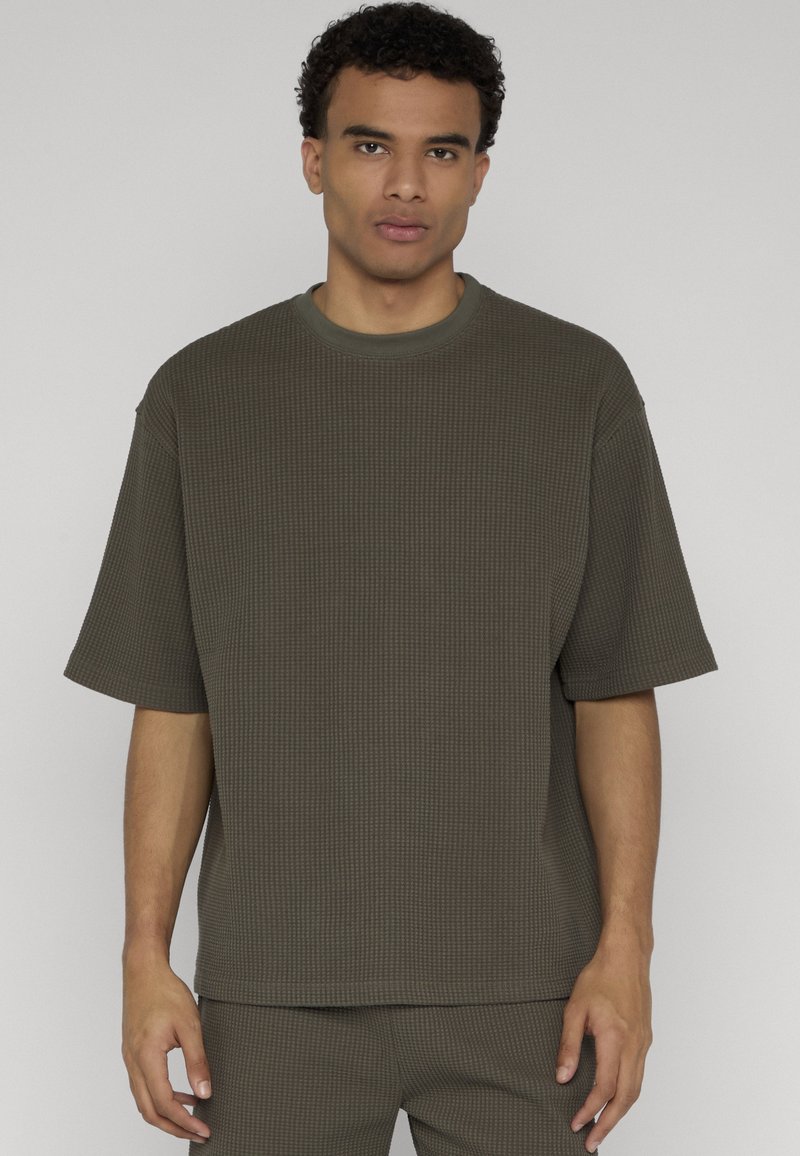 Homme aux cheveux courts et bouclés portant une chemise à manches courtes en tricot gaufré olive surdimensionnée et un pantalon assorti, debout devant un fond uni.