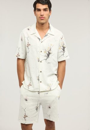 Giovane uomo che indossa una camicia a maniche corte con bottoni e pantaloncini abbinati con motivi floreali leggeri e crepati. Mani nelle tasche.