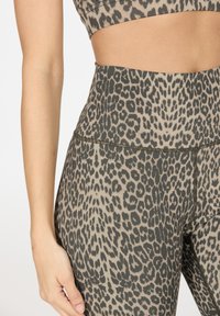 Leopardprint hoge taille legging en bijpassende sportbeha, met een cropped middengedeelte en één arm ontspannen aan de zijkant.