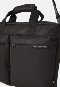 Sort nylon bærepose til laptop med to forlommer, top-håndtag og mærket rem. Indeholder et logo-label og en diskret struktur.