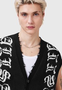 Melns adīts veste ar baltu grafisku tekstu, kam ir V veida kakls, poga priekšpusē un nodilušas malas. Valkāts virs baltas tanktopas.