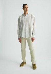 Camisa de botones a rayas en tonos claros, con un ajuste relajado, mangas largas y textura suave; combinada con pantalones de color claro y zapatos marrones.