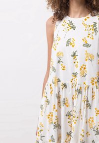 Model iført en ærmeløs hvid kjole med gule blomster mønstre, der har en tætsiddende bodice og et lagdelt skørt. Baggrunden er lys.