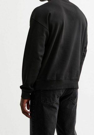 Černá mikina s volnějším střihem, žebrovanými rukávy a lemem, vyrobená z hladké tkaniny, ve spojení s tmavými džínami, pohled zezadu.