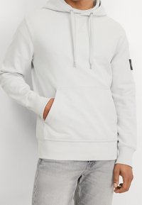 Ljusgrå huvtröja med känguruficka, dragsnodd på huvan, ribbade ärmslut och nederkant, samt en liten svart logotyp på ärmen.