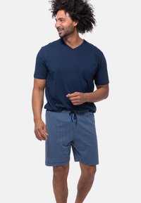 Mann mit lockigem Haar, der ein navyblaues V-Ausschnitt-Shirt und karierte Shorts trägt, lächelt und gestikuliert mit seiner Hand. Weißer Hintergrund.