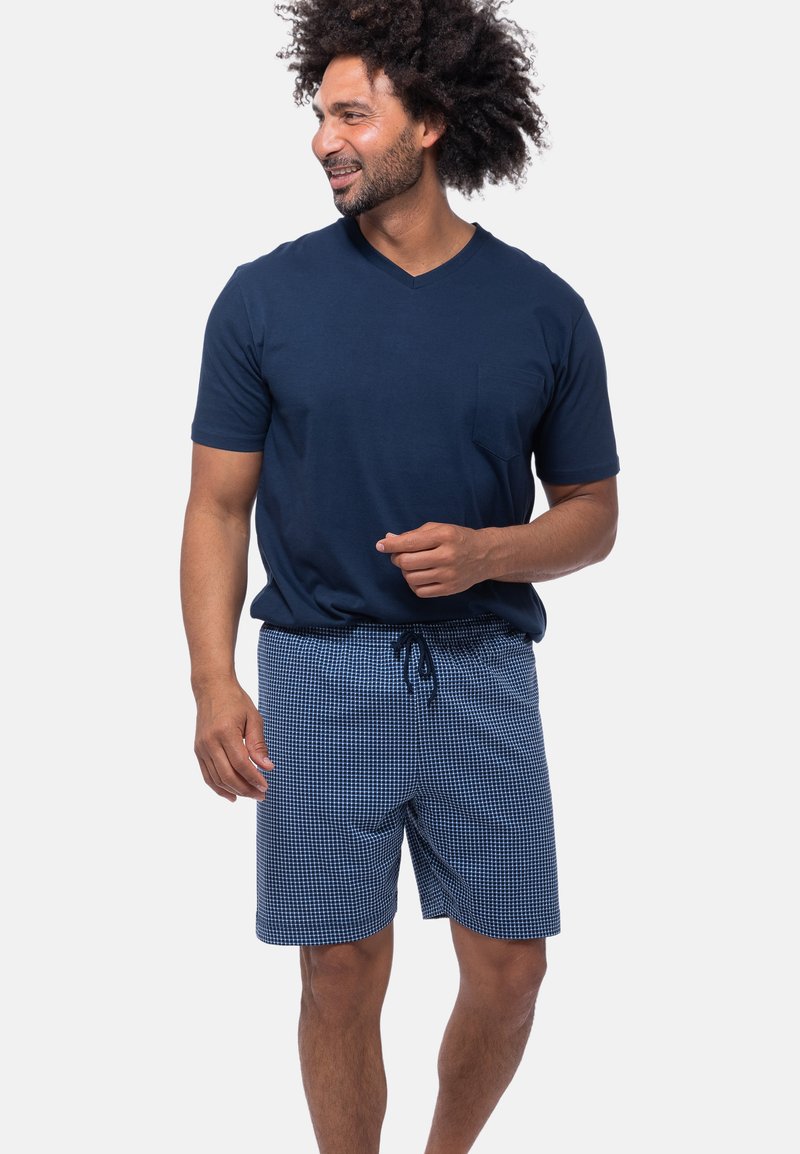 Mann mit lockigem Haar, der ein navyblaues V-Ausschnitt-Shirt und karierte Shorts trägt, lächelt und gestikuliert mit seiner Hand. Weißer Hintergrund.