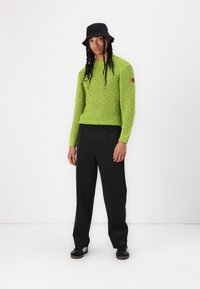 Suéter de malha em verde limão brilhante com um corte cropped, combinado com calças largas pretas e um chapéu tipo balde preto. Ténis pretos completam o visual.