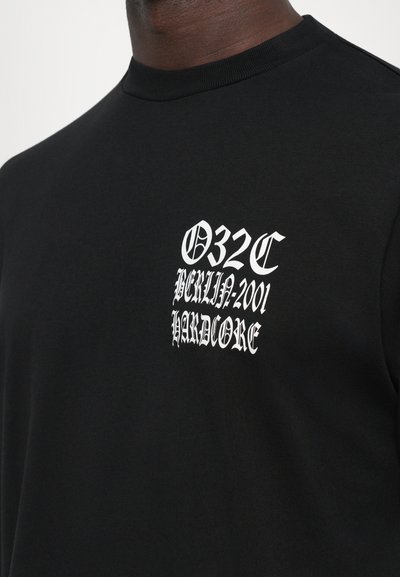 Nahaufnahme eines schwarzen Shirts mit weißem Gothic-Text auf der Brust, der "032C Berlin 2001 Hardcore" liest.