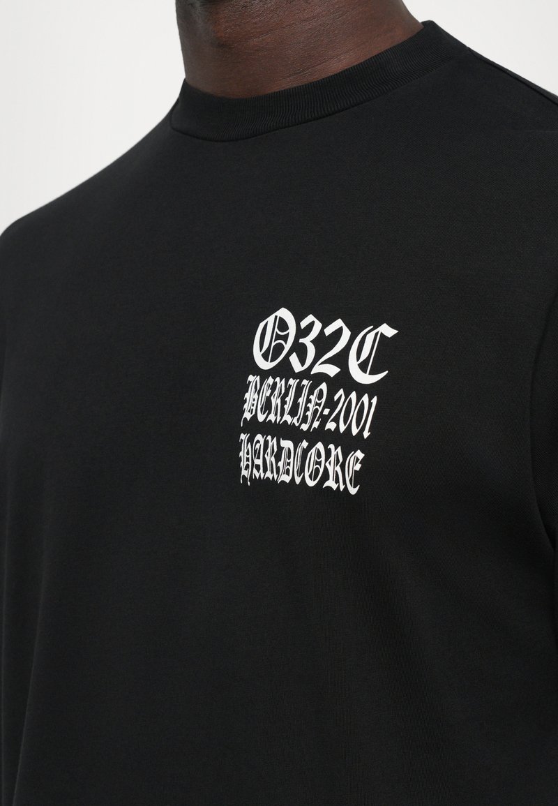 Gros plan sur une chemise noire avec un texte gothique blanc sur la poitrine indiquant "032C Berlin 2001 Hardcore."