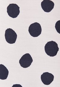 Tissu blanc avec des pois noirs irréguliers, montrant un tissage texturé et des formes et tailles de pois variées.