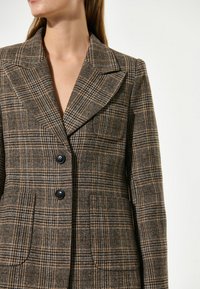 Blazer a quadri marroni con collo a rovescio, due grandi bottoni e tasche frontali, realizzato in tessuto testurizzato con linee a contrasto.