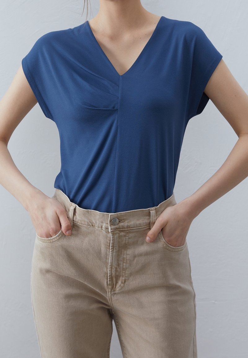 Navy blue V-neck top with short sleeves and a front pocket, paired with beige high-waisted pants. Smooth fabric, relaxed fit, minimal design.