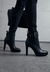 Czarne skórzane botki za kostkę na wysokim obcasie, z bocznym zamkiem błyskawicznym i detalem w postaci paska. Noszone z ciemnymi skinny jeansami na szarym tle.