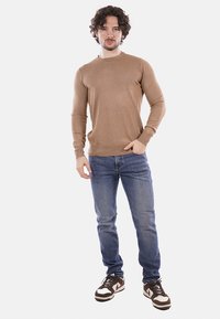 Maglione a maglia marrone chiaro con collo tondo, abbinato a jeans in denim blu e sneakers marroni e bianche. Il modello si trova di fronte a uno sfondo semplice.