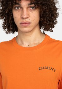 Camiseta de cuello redondo naranja hecha de algodón suave, que presenta un logotipo negro de "ELEMENT" bordado en el pecho, tela lisa con textura suave.