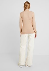Femme aux longs cheveux tressés portant un pull à col roulé beige, un pantalon large en velours côtelé crème et des baskets blanches, debout de dos.
