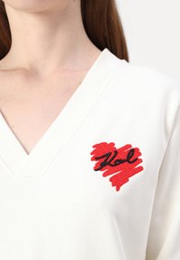 Kreminė V-kaklo megztinis su raudonu širdies dizainu, šepetėliu detalėmis ir juodu siuvinėjimu "Karl" kairėje krūtinės pusėje.