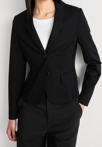 Zwarte op maat gemaakte blazer met een getailleerd ontwerp, met schouderflappen, twee knopen en voorzakken, gemaakt van gladde stof met een matte afwerking.