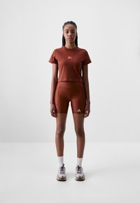 Rust-colored athletic top and shorts set, featuring a logo on the shirt and a triangle logo on the shorts. Worn with white socks and shoes.