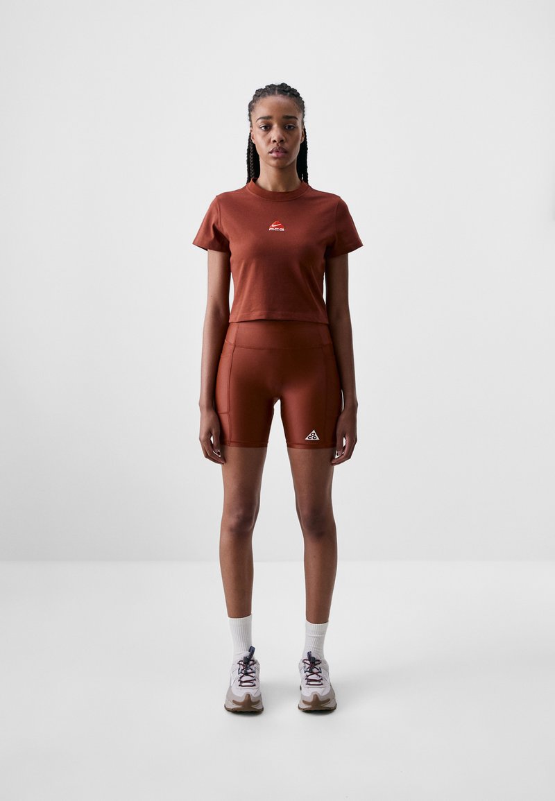 Rust-colored athletic top and shorts set, featuring a logo on the shirt and a triangle logo on the shorts. Worn with white socks and shoes.