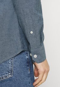 Blauwe chambray shirt met een knoopsluiting aan de manchetten, met een lichte textuur en een afgeronde zoom. Draagt bij een denimjeans, die stikseldetails toont.