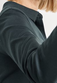 Mörkgrön skjorta med krag-nacke, som har en slät yta och figursydda ärmar, med tydliga sömdetaljer nära axeln.