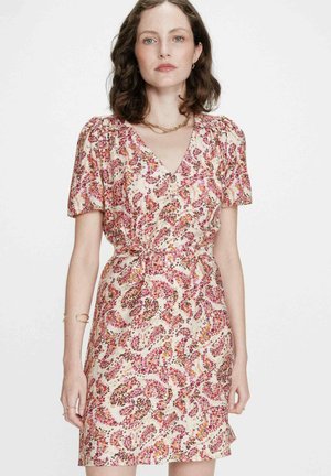 Femme portant une robe florale à manches courtes et col en V avec des motifs roses, orange et jaunes, accessoirisée de bijoux en or sur fond blanc.