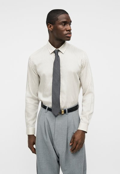 Άνδρας με λευκό πουκάμισο με ρίγες, γκρι γραβάτα, γκρι πλισέ παντελόνι και μαύρη ζώνη, που κοιτάζει πλάγια μπροστά σε απλό φόντο.