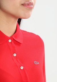 Žena v červeném polo tričku se třemi bílými knoflíky a malým krokodýlím logem na levé straně hrudi.