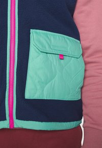 Marineblaue Fleece-Weste mit einer gesteppten mintgrünen Tasche. Ausgestattet mit einem rosa Rei�ßverschluss und strukturierten Akzenten. Auch über einem hellrosa Oberteil getragen.