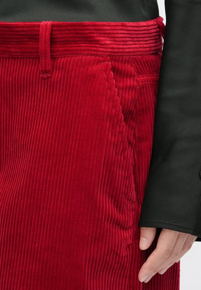 Red corduroy skirt with vertical ribbed texture, featuring a waistband and side pocket. Accompanied by a black smooth-sleeved top.
