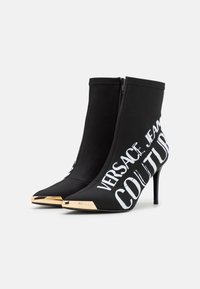 Czarne botki za kostkę z materiałową cholewką, szpilką, spiczastym noskiem i złotymi detalami. Na obuwiu znajduje się biały nadrukowany tekst "Versace Jeans Couture".