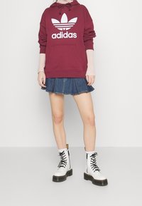 Felpa bordeaux con logo Adidas bianco, gonna di jeans plissettata e stivali bianchi con lacci, suola nera e tab giallo sul tallone.