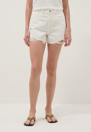 Person trägt weiße, ausgewaschene Jeansshorts und olivgrüne Sandalen mit Metallösen, stehend vor einem schlichten hellen Hintergrund.