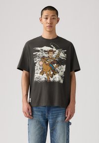 Camiseta de algodón gris con un estampado gráfico de un vaquero de dibujos animados y un caballo, con mangas cortas y un ajuste relajado. Jeans de mezclilla visibles debajo.