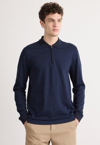 Polo de manga longa azul marinho com gola com fecho, apresentando um padrão texturizado e punhos elásticos, combinado com calças de cor clara.