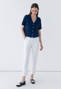 Marinblå kortärmad cardigan med guldbutiker, kombinerad med vita skräddarsydda byxor och svarta loafers med gröna accenter. Släta texturer och modern passform.