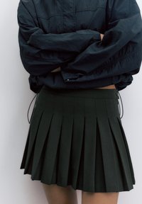 Person iført en plisseret sort mininederdel og en sort langærmet skjorte med krydsede arme foran en ensfarvet baggrund.