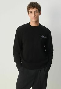 Sort strikket sweater med rund halsudskæring, ribbede manchetter og kant, prydet med hvid broderet tekst på brystet. Blød tekstur og tætsiddende design.