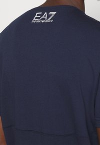 Navy cotton T-shirt featuring a round neckline, short sleeves, and a white EA7 Emporio Armani logo on the back. Smooth texture, simple design.
