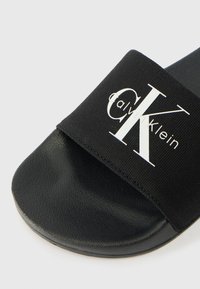 Μαύρη σανδάλι με φαρδύ ύφασμα που φέρει το λευκό λογότυπο "Calvin Klein". Υφή στη σόλα, λείος σχεδιασμός και μινιμαλιστική αισθητική.