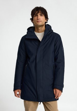 Hombre que lleva una chaqueta de invierno azul marino con capucha y pantalones beige, de pie frente a un fondo blanco liso.